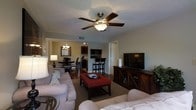 This single-floor residence offers a thoughtfully designed open-concept layout that seamlessly connects the main living, dining, and kitchen areas. The living room is comfortably furnished with neutral-toned sofas surrounding a bold red upholstered ottoman that serves as a focal point, complemented by a media console with a large flat-screen television. A ceiling fan with an integrated light fixture provides both comfort and illumination, while a textured ceiling adds subtle architectural character throughout.

The dining area sits adjacent to the living space and features a dark wood dining table with matching chairs beneath an elegant chandelier, creating a warm and inviting atmosphere. A large mirror on the wall enhances the sense of depth in this area.

The kitchen is equipped with dark wood cabinetry, black appliances including a built-in microwave, and a raised bar counter with bar stools — ideal for casual dining or entertaining. The space benefits from consistent flooring that flows throughout, tying all areas together cohesively.

Overall, the space presents a harmonious blend of dark wood tones and light, neutral furnishings against soft gray-green walls, creating a balanced and welcoming interior that is both functional and aesthetically cohesive.