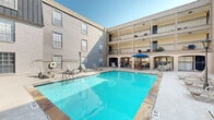 Located along the vibrant Westheimer Road corridor in Houston, Texas, this outdoor amenity space offers a well-appointed courtyard experience designed for relaxation and leisure. At the heart of the space is a generously sized swimming pool with clear turquoise water, framed by a stone-tiled surround and ample poolside deck area outfitted with lounge chairs for sunbathing. A life preserver ring is stationed nearby, underscoring the safety-conscious design of the pool area. The courtyard is enclosed on multiple sides by a multi-story building featuring covered upper-level balconies with wrought iron railings, siding-clad facades, and large windows that overlook the outdoor space below. Adjacent to the pool, a spacious open-air patio extends the outdoor living area and includes a wooden pergola providing shaded seating with lounge chairs positioned beneath it. A dining area furnished with a round table, wooden chairs, and a bold blue patio umbrella offers a comfortable spot for outdoor meals. White-painted brick walls lend a classic character to portions of the exterior, while classic lantern-style light posts add a charming architectural touch. The open sky above ensures abundant natural light throughout the day, creating a bright and inviting atmosphere ideal for outdoor living.