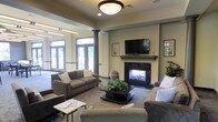 This single-floor space is designed as an inviting communal lounge that balances comfort and functionality. The room centers around a classic fireplace with a dark surround and a traditional mantel, above which a wall-mounted flat-screen television is positioned for easy viewing. Decorative framed artwork flanks the fireplace, adding a refined touch to the warm, neutral color palette of the walls.

The seating arrangement is generous and thoughtfully laid out, featuring plush upholstered sofas and armchairs in muted earth tones, oriented around a dark wood coffee table adorned with a lush green plant. A textured area rug anchors the seating area and adds warmth underfoot. A secondary small side table provides additional surface space for guests.

Architectural character is conveyed through prominent dark decorative columns that frame the transition between areas, adding a sense of structure and elegance to the open layout. Flush-mount and recessed ceiling lighting ensures the space is well-lit throughout.

Natural light pours in through large glass-paned French doors along the far wall, offering a view of a verdant outdoor area and creating a bright, airy atmosphere. The overall ambiance is welcoming and polished, making this lounge well-suited for social gatherings, relaxation, or casual meetings.