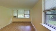 This single-floor space presents a straightforward and functional interior highlighted by warm hardwood-style flooring that runs throughout the main living area. The room is characterized by a bank of windows fitted with horizontal blinds, allowing ample natural light to fill the space while offering pleasant views of mature trees and outdoor greenery beyond. A window-unit air conditioner is installed in one of the lower window panels, providing climate control during warmer months. Baseboard heating runs along the perimeter walls, ensuring warmth throughout the cooler seasons. The walls are finished in a soft, neutral yellow-green tone that contributes to a bright and airy atmosphere. A partial wall or built-in ledge is visible along one side of the room, which may serve as a functional divider or display surface. The overall layout feels open and uncluttered, with clean lines and minimal architectural detailing. The combination of natural light, outdoor views, and practical heating and cooling systems makes this a comfortable and livable space well-suited for everyday use.