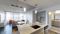 This single-floor space showcases a seamless open-concept layout that integrates the kitchen, dining, and living areas into one cohesive environment. The kitchen is anchored by a large island with a sleek white countertop and an undermount sink, complemented by a brushed chrome faucet. Overhead, cylindrical pendant lights hang from the exposed concrete ceiling, adding an industrial edge that contrasts beautifully with the clean, modern aesthetic. The cabinetry is finished in a smooth matte grey, offering ample storage with a built-in wine rack and display space for glassware.

The dining area features a round white table with matching chairs, positioned near floor-to-ceiling windows that invite generous natural light and offer views of the surrounding cityscape. Track lighting mounted along the exposed concrete ceiling provides additional illumination throughout the space. Warm hardwood flooring grounds the interior, adding texture and contrast to the otherwise cool palette of whites and greys. The overall ambiance is sleek and contemporary, blending industrial architectural elements with refined, minimalist finishes for a polished urban feel.