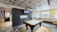 This thoughtfully designed single-floor amenity space blends industrial character with upscale comfort across several distinct zones. The standout feature is the game and entertainment area, anchored by a full-size billiards table set atop a patterned area rug. A dramatic dark-paneled accent wall serves as the backdrop for a wall-mounted flat-screen TV and a sleek linear fireplace with a glass surround, creating a striking focal point. Throughout the space, ornate embossed tin ceilings add a vintage architectural detail that contrasts beautifully with the contemporary herringbone-patterned luxury vinyl flooring below. A reclaimed wood accent column adds warmth and texture, reinforcing the space's eclectic, industrial-chic aesthetic. Track lighting and statement pendant fixtures provide layered illumination throughout. The kitchenette is outfitted with white shaker cabinetry, dark granite countertops, a stainless steel farmhouse sink, open wood shelving, and stainless steel appliances, all complemented by classic white subway tile. A separate lounge area offers a tufted leather Chesterfield sofa, patterned accent chairs, and a sleek coffee table — ideal for relaxed gatherings. Natural light filters in through multiple windows fitted with horizontal blinds, enhancing the overall ambiance of the space.