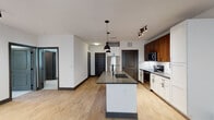 This single-floor unit showcases a thoughtfully designed open-concept layout where the kitchen and living area flow seamlessly together. Light wood-tone flooring runs throughout the main living spaces, complemented by crisp white walls and dark baseboards that create a clean, contemporary contrast.

The kitchen is a standout feature, combining white shaker-style lower cabinets with rich walnut-toned upper cabinetry. A white subway tile backsplash adds texture, while stainless steel appliances — including a refrigerator, range, and built-in microwave — provide a polished finish. A central island with a dark countertop and an undermount sink serves as both a functional workspace and a visual anchor, accented by matte black pendant lighting overhead. Track lighting along the kitchen perimeter offers additional illumination.

The open living area benefits from natural light through a window with exterior views and features a ceiling fan. Bifold doors lead to an additional room or closet space, adding versatility.

The bedroom is finished with plush neutral carpet and includes a ceiling fan, a window with outdoor views, and ample wall space. The bathroom presents a double-sink vanity with a dark countertop, white shaker cabinets, backlit rectangular mirrors, a soaking tub, and a separate tiled shower with a glass enclosure.