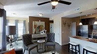 This single-story space showcases a thoughtfully designed open-concept layout that seamlessly connects the living area to adjacent spaces. The living area is anchored by rich, dark hardwood flooring that contrasts elegantly with the warm beige and bold chocolate-brown accent wall, which serves as a striking backdrop for colorful wall art. A ceiling fan with integrated lighting fixture adds both functionality and a finishing touch to the space overhead.

The seating area features tufted gray accent chairs paired with a round side table, creating an intimate conversational arrangement. A low-profile console table against the accent wall provides additional display space for decorative objects. Geometric metal wall sculptures add a contemporary artistic element to the neutral-toned walls.

Natural light filters in through a window dressed with sheer curtains, brightening the open interior. A glimpse of the kitchen area to the right reveals a raised countertop with bar seating, suggesting a practical and social dining option. Multiple doorways visible throughout indicate well-connected rooms, contributing to a functional and cohesive floor plan with a modern, polished ambiance.
