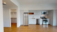 This single-floor unit showcases a clean, contemporary design with a cohesive white-and-neutral palette throughout. The open kitchen is a standout feature, outfitted with flat-front, light grey cabinetry extending to the ceiling for substantial storage. A full suite of stainless steel appliances includes a gas range, over-the-range microwave, dishwasher, and a full-size refrigerator. A warm wood-toned open shelf adds a subtle accent against the otherwise crisp white subway tile backsplash and light countertops. Hardwood-style flooring runs throughout the main living areas, contributing to a bright and cohesive aesthetic.

The bathroom features a full soaking tub and shower combination dressed in large-format marble-look tile, complemented by grey floor tile and a clean white finish. A spacious walk-in closet provides generous storage with multiple hanging rods and shelving on three walls, finished with the same warm hardwood-style flooring. The overall layout, as seen in the floor plan view, is efficiently organized, connecting the kitchen, bathroom, and closet in a logical flow that maximizes functionality within the space.