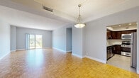 This single-floor residence offers a thoughtfully designed layout with a variety of distinct living spaces connected by open sightlines and consistent finishes throughout. The main living and dining areas are unified by warm parquet hardwood flooring arranged in a classic basket-weave pattern, complemented by soft grey walls, white baseboards, and a textured ceiling. Pendant light fixtures with brushed metal finishes anchor the dining zone, while a tray ceiling detail adds subtle architectural interest. Large sliding glass doors lead to a private balcony enclosed by ornate metal railings, offering elevated views of a lush, tree-lined landscape — particularly striking during autumn foliage. The kitchen is well-equipped, featuring rich dark wood cabinetry, granite countertops with a subtle speckled finish, a white subway tile backsplash, and stainless steel appliances including a gas range and over-the-range microwave. The bedroom is finished with plush grey carpeting, providing a quieter, softer retreat. The bathroom includes a granite-topped vanity with white cabinetry, an undermount sink, and a glass-enclosed shower, presenting a clean and functional design. Throughout, the space maintains a neutral, versatile palette suited to a range of interior styles.
