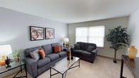 This single-story space centers around a thoughtfully arranged living room that balances comfort and contemporary style. The room is finished with soft, neutral-toned carpet and light gray walls, creating a calm and inviting atmosphere. A large window fitted with white horizontal blinds allows natural light to filter in, brightening the space while offering privacy.

The living area is furnished with a matching set of charcoal gray upholstered sofas — a full-length couch and a loveseat — accented with warm rust and orange throw pillows that add a touch of color contrast. A sleek glass-top coffee table with a minimalist metal frame serves as the focal point of the seating arrangement, complemented by a matching glass side table positioned between the sofas.

Decorative elements include a pair of framed abstract wall art pieces in coordinating earth tones, a classic table lamp, and a small potted plant in the corner, contributing to the overall warmth of the space. The clean lines, cohesive color palette, and functional layout make this living room both practical and visually appealing.