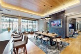 This expansive amenity lounge is designed with a modern, upscale aesthetic that balances comfort and functionality across several well-defined zones. Light hardwood flooring flows continuously throughout, unifying the space while complementing the varied design elements found in each area.

The entry foyer is anchored by bold navy accent walls, a marble-clad feature with pendant lighting, and steel-framed glass doors that provide a sense of transparency and openness. Rounded gold upholstered chairs add a welcoming touch near the entrance.

Moving deeper into the space, a lounge area features a dark wood slatted partition wall with a mounted flat-screen television, flanked by clusters of gold boucle chairs and a circular side table. An adjacent sunlit area offers leather lounge chairs positioned along floor-to-ceiling glass walls that frame views of an outdoor pool courtyard.

A dedicated dining and gathering zone showcases a warm wood-plank ceiling with integrated cove lighting, a live-edge communal table beneath statement pendant fixtures, and a dark stone accent wall with another mounted screen. A full wet bar with a sculpted concrete island, stone backsplash, and dark cabinetry serves as a central entertaining hub. Additional seating areas with a fireplace and billiards table complete the layout.