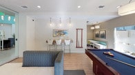 This well-appointed single-floor amenity space blends modern design with a variety of recreational and functional areas. At the heart of the main lounge is a full-size billiards table, positioned beneath a statement pendant light and surrounded by warm hardwood flooring. Teal accent walls add a vibrant pop of color, complementing the neutral tones throughout. Large windows and glass doors flood the space with natural light and provide direct access to an outdoor patio, where additional seating and greenery are visible.

A comfortable lounge area features a wall-mounted flat-screen television set against warm wood paneling, with a drum pendant light overhead and a mix of seating options including a sofa and patterned loveseat. A glass-enclosed conference or meeting room is accessible from the lounge, offering a private yet visually connected workspace complete with pendant lighting and a dedicated table with chairs.

The space also includes a bar-height counter with stools beneath a row of Edison-style pendant lights, ideal for casual gatherings. The kitchenette is efficiently designed with wood cabinetry, light countertops, a stainless steel refrigerator, and a sink — accompanied by a dining table and cheerful green chairs. Together, these areas create a versatile, community-oriented environment suited for socializing, working, and recreation.