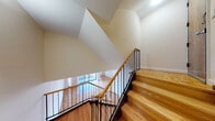 This thoughtfully designed residence offers a harmonious blend of classic finishes and functional layout across its split-level floor plan. The centerpiece of the living area is the warm, intricately patterned parquet wood flooring that extends throughout, lending a timeless character to the space.

The main living area is generously proportioned, bathed in natural light through large picture windows that frame views of the surrounding urban neighborhood. Floor-to-ceiling black-framed sliding glass doors open to a private balcony, seamlessly connecting the interior to the outdoors and further enhancing the sense of openness.

A striking staircase with natural wood treads and a sleek black iron railing with a wooden handrail serves as an architectural focal point, connecting the living level to an upper landing. The angular, vaulted ceiling above the stairwell adds visual drama and a sense of height to the space.

The kitchen is efficiently laid out with white raised-panel cabinetry, dark granite-style countertops, white appliances including a gas range and dishwasher, and durable slate-toned flooring. The bathroom is cleanly finished with white subway tile surrounding the bathtub and shower, dark mosaic tile flooring, and a mirrored medicine cabinet. An additional room with its own sliding glass balcony access offers flexible use as a bedroom or study.