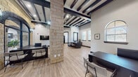 This single-floor commercial space makes a striking architectural statement from the moment of entry, where a grand arched glass doorway framed by natural limestone masonry sets the tone for the interior. The facade's stacked stone columns and rounded archways establish a refined yet rustic character that continues throughout the space.

Inside, the open layout is defined by soaring ceilings adorned with bold, dark-stained exposed wood beams that contrast sharply against crisp white walls. Structural limestone columns rise from the light wood-look flooring, reinforcing the building's distinctive material palette while dividing the space into functional zones without the need for full walls. Dark-framed windows fitted with wood blinds line the perimeter, allowing ample natural light to filter through while maintaining privacy.

Multiple workstation areas are arranged throughout the open floor plan, each equipped with dark-finished desks and contemporary seating. Black-trimmed doorways lead to private offices or ancillary rooms, providing separation when needed. Arched transom windows add architectural interest and draw the eye upward. A wall-mounted flat-screen display, decorative framed artwork, and carefully placed greenery contribute to a polished, professional ambiance throughout.