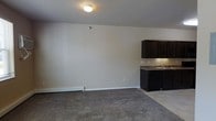 This single-floor space offers a straightforward, functional layout that balances living and kitchen areas within an open floor plan. The main living area is finished with wall-to-wall carpet in a neutral gray tone, creating a soft, comfortable underfoot feel that complements the light beige walls throughout. A flush-mount ceiling light provides overhead illumination, while a baseboard heating unit runs along the exterior wall, ensuring warmth throughout the space.

The kitchen area transitions seamlessly from the carpeted living space onto tile flooring, providing a practical and clean delineation between the two zones. Dark espresso-toned cabinetry lines the kitchen, offering ample storage with a rich, bold contrast against the lighter walls. The countertops feature a granite-look surface in muted earth tones, paired with a stainless steel sink. The overall design aesthetic is clean and understated, with a neutral color palette that lends itself well to a variety of furnishing styles.

The space is practical and efficiently arranged, making the most of the available footprint. The minimal detailing and open feel give the interior a versatile, move-in-ready character suitable for a range of uses.