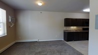 This single-floor space offers a straightforward, functional layout that balances living and kitchen areas within an open floor plan. The main living area is finished with wall-to-wall carpet in a neutral gray tone, creating a soft, comfortable underfoot feel that complements the light beige walls throughout. A flush-mount ceiling light provides overhead illumination, while a baseboard heating unit runs along the exterior wall, ensuring warmth throughout the space.

The kitchen area transitions seamlessly from the carpeted living space onto tile flooring, providing a practical and clean delineation between the two zones. Dark espresso-toned cabinetry lines the kitchen, offering ample storage with a rich, bold contrast against the lighter walls. The countertops feature a granite-look surface in muted earth tones, paired with a stainless steel sink. The overall design aesthetic is clean and understated, with a neutral color palette that lends itself well to a variety of furnishing styles.

The space is practical and efficiently arranged, making the most of the available footprint. The minimal detailing and open feel give the interior a versatile, move-in-ready character suitable for a range of uses.