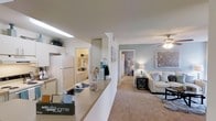 This single-floor residence offers a thoughtfully designed open-concept layout that seamlessly connects the kitchen and living areas. The kitchen features a peninsula-style counter with ample workspace, white cabinetry extending to the ceiling for generous storage, and full-size appliances including a refrigerator. A skylight above the kitchen brings in additional brightness, complementing the warm overhead lighting throughout. The adjoining living area is anchored by soft, neutral-toned walls in a calming blue-grey hue, wall-to-wall carpeting, and a ceiling fan that adds both style and functionality. The space is furnished with a light-colored sofa accented by blue and white throw pillows, creating a cohesive and relaxing atmosphere. A glimpse down the hallway reveals access to additional rooms, including what appears to be a laundry area with stacked or side-by-side units. The overall ambiance is clean, bright, and welcoming, with a neutral color palette that lends itself well to a variety of décor styles. The open sightlines between the kitchen and living room enhance the sense of spaciousness, making this a practical and comfortable single-floor home.