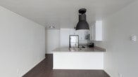 This single-floor space showcases a clean, contemporary design with an open-concept layout that maximizes flow and functionality. The kitchen serves as the centerpiece of the space, featuring crisp white shaker-style cabinetry that extends to the upper walls, providing ample storage while reinforcing the bright, minimalist aesthetic. A peninsula island with a neutral-toned countertop offers additional workspace and doubles as a casual dining or gathering area, complete with an integrated sink and modern fixtures. A bold, oversized black industrial-style pendant light hangs above the island, creating a striking contrast against the white surroundings and serving as a standout design feature. Stainless steel appliances, including a full-size refrigerator, microwave, and range, lend a polished, modern touch to the kitchen suite. Dark hardwood flooring runs throughout the space, grounding the light palette and adding warmth and depth to the overall ambiance. The walls are finished in a clean, bright white, enhancing the sense of openness and allowing natural and artificial light to reflect evenly throughout. The space presents a cohesive, well-designed environment that balances modern style with practical living.
