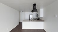 This single-floor space showcases a clean, contemporary design with an open-concept layout that maximizes flow and functionality. The kitchen serves as the centerpiece of the space, featuring crisp white shaker-style cabinetry that extends to the upper walls, providing ample storage while reinforcing the bright, minimalist aesthetic. A peninsula island with a neutral-toned countertop offers additional workspace and doubles as a casual dining or gathering area, complete with an integrated sink and modern fixtures. A bold, oversized black industrial-style pendant light hangs above the island, creating a striking contrast against the white surroundings and serving as a standout design feature. Stainless steel appliances, including a full-size refrigerator, microwave, and range, lend a polished, modern touch to the kitchen suite. Dark hardwood flooring runs throughout the space, grounding the light palette and adding warmth and depth to the overall ambiance. The walls are finished in a clean, bright white, enhancing the sense of openness and allowing natural and artificial light to reflect evenly throughout. The space presents a cohesive, well-designed environment that balances modern style with practical living.