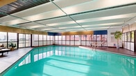 Located at 1212 Avenue des Pins Ouest in Montréal, this generously sized indoor pool facility offers a refined aquatic experience within a single, open-plan level. The centerpiece is a large, rectangular lap pool with vivid turquoise waters, framed by wide-format floor tiles that extend along the perimeter deck. The architectural design features a distinctive beamed ceiling with alternating light and dark paneling accented by teal and navy tones, giving the space a structured yet vibrant character. Recessed and linear lighting supplements the abundant natural light that streams in through an expansive grid of floor-to-ceiling windows lining multiple walls, offering views of the surrounding Montréal skyline and cityscape. The surrounding deck is furnished with contemporary lounge seating, including a modern sofa set with a low coffee table and stylish hanging egg chairs, providing comfortable areas to relax before or after a swim. Glass-paneled entry doors allow smooth access to and from adjoining areas. A dedicated bench and pool access ladder are positioned poolside for convenience. The overall ambiance is clean, modern, and inviting, blending functional aquatic infrastructure with thoughtfully curated lounge elements to create a well-rounded recreational space.
