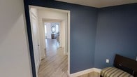 This single-floor space presents a well-coordinated interior design anchored by striking navy blue accent walls that create a bold yet refined atmosphere. The hallway serves as a central connector, drawing the eye toward the living areas beyond through a series of clean, white-trimmed doorways and archways that add architectural interest and a sense of depth.

The light wood-look flooring runs seamlessly throughout, lending warmth and continuity to the space. Bright white baseboards and door casings provide crisp contrast against the deep blue walls, reinforcing a polished, modern aesthetic. The corridor layout is open and unobstructed, allowing natural light from the adjoining rooms to filter through and brighten the passageway.

Beyond the hallway, a living or sitting area is visible, furnished with tasteful pieces including a decorative round mirror that serves as a focal point on the wall, complemented by a sideboard or credenza in a warm wood tone. The overall ambiance is calm and sophisticated, blending contemporary color choices with classic architectural details. The thoughtful use of color, light, and consistent materials throughout creates a cohesive, inviting environment that feels both stylish and comfortable.