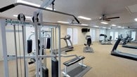 Located at 700 N Albert Pike Ave in Fort Smith, Arkansas, this single-floor fitness center offers a well-equipped space designed to support a comprehensive workout routine. The room features a multi-station cable weight machine with an attached bench, providing options for a wide range of strength training exercises. Cardio equipment includes a treadmill and a stationary bike, both positioned on protective floor mats. A full-length mirror spans one wall, enhancing the sense of space and allowing users to monitor their form during workouts. A wall-mounted television adds a layer of entertainment for those looking to stay engaged during longer sessions. The space is finished with a textured ceiling and lit by recessed fluorescent panel lighting, supplemented by ceiling fans that help maintain air circulation throughout the room. The neutral-toned walls and speckled carpeted flooring contribute to a clean, functional aesthetic. The overall layout is open and organized, making it easy to navigate between equipment while maintaining a comfortable workout environment.