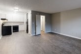 This single-floor unit offers a clean, functional layout designed for comfortable everyday living. The main living area is finished with durable wood-style flooring in a warm medium-tone, complemented by neutral gray walls and crisp white baseboards that create a fresh, modern aesthetic. A textured ceiling runs throughout the space, adding subtle visual character. The open layout connects seamlessly to the kitchen, which features white cabinetry with ample upper and lower storage, light-toned countertops, and a full suite of black appliances including a range, dishwasher, and refrigerator. Light wood-style flooring in the kitchen provides a clean contrast to the darker tones in the living area. A dedicated bedroom offers a quiet retreat, featuring a wall-mounted air conditioning unit and a window that allows natural light to filter in. The bathroom is practical and well-appointed, with extended granite-style countertop space, a large wall mirror, a single sink, and a tiled tub-shower combination. Throughout the unit, the consistent use of neutral tones, white trim, and hard-surface flooring creates a cohesive and easy-to-maintain living environment.