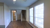 This two-story home offers a practical and comfortable layout with notable design details throughout. The main level features rich, dark hardwood flooring that anchors the open living area, complemented by neutral-toned walls that create a clean, versatile backdrop. Large windows along one wall allow abundant natural light to fill the space, enhancing the open and airy feel of the room. A ceiling fan is installed overhead, contributing to comfort year-round. Carpeted stairs with a white railing lead to the second floor, providing a clear visual transition between levels. Adjacent to the living area, a doorway opens into a well-lit bathroom or utility space, visible with track lighting, a vanity, and mirror, adding convenience to the main floor. The textured ceiling adds a subtle design element consistent throughout the space. The overall ambiance is neutral and move-in ready, with a straightforward layout that balances functionality and comfort across both floors.