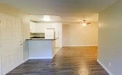 This single-floor space features an open-concept design that seamlessly connects the kitchen and living areas, creating a cohesive and functional layout. The living area is finished with warm-toned, hardwood-style laminate flooring that extends throughout, adding a sense of continuity and visual warmth. A ceiling fan with an integrated light fixture is centrally positioned in the living space, providing both ventilation and ambient lighting.

The kitchen is defined by clean white cabinetry that reaches toward the ceiling, offering generous storage capacity. A raised breakfast bar with a dark countertop serves as a natural dividing element between the kitchen and the living area, providing additional seating or workspace. White appliances, including a refrigerator, complement the bright, neutral palette of the kitchen.

The walls throughout are finished in a soft, neutral tone that enhances the sense of openness and allows natural light to reflect evenly across the space. White baseboards provide clean, crisp definition along the floors. Overall, the space presents a straightforward, well-maintained interior with a practical and welcoming ambiance suited for everyday living.