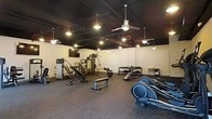 This fitness center offers a well-rounded workout environment designed to accommodate a range of exercise needs. The space features a dark, exposed ceiling with industrial-style ductwork, giving it a modern, urban aesthetic. Ceiling fans and pendant lighting are suspended throughout, providing both air circulation and warm, focused illumination across the workout floor.

The flooring consists of durable rubber gym matting, which provides cushioning and traction ideal for high-activity use. Equipment is thoughtfully arranged to maximize usable space, including a multi-station cable machine, incline and flat benches, a rowing machine, and elliptical trainers. A dumbbell rack stocked with a variety of weights is positioned along one wall, complemented by a small utility table nearby.

A flat-screen television is mounted on the wall, adding an entertainment option for users during their workouts. The light-colored walls contrast with the dark ceiling, keeping the space feeling open and bright despite the moody overhead aesthetic. Overall, the fitness center presents a functional and well-maintained environment suited for both strength training and cardiovascular exercise.