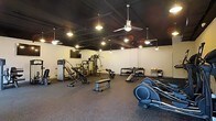 This fitness center offers a well-rounded workout environment designed to accommodate a range of exercise needs. The space features a dark, exposed ceiling with industrial-style ductwork, giving it a modern, urban aesthetic. Ceiling fans and pendant lighting are suspended throughout, providing both air circulation and warm, focused illumination across the workout floor.

The flooring consists of durable rubber gym matting, which provides cushioning and traction ideal for high-activity use. Equipment is thoughtfully arranged to maximize usable space, including a multi-station cable machine, incline and flat benches, a rowing machine, and elliptical trainers. A dumbbell rack stocked with a variety of weights is positioned along one wall, complemented by a small utility table nearby.

A flat-screen television is mounted on the wall, adding an entertainment option for users during their workouts. The light-colored walls contrast with the dark ceiling, keeping the space feeling open and bright despite the moody overhead aesthetic. Overall, the fitness center presents a functional and well-maintained environment suited for both strength training and cardiovascular exercise.