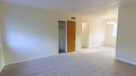 This single-floor residential space offers a straightforward and functional layout designed for comfortable everyday living. The interior is characterized by neutral, light-toned walls that create a bright and airy atmosphere throughout. The bedroom area is generously proportioned, featuring wall-to-wall carpeting in a warm beige tone that adds softness underfoot. A built-in closet with a classic wood-paneled door provides convenient storage, complete with interior shelving and a hanging rod for organized use. A textured ceiling adds a subtle architectural detail to the room. An open doorway connects the bedroom to an adjacent space, suggesting a well-connected floor plan that allows for easy movement between rooms. The overall ambiance of the space is clean, understated, and move-in ready, with a simple aesthetic that serves as a neutral canvas for personalization. Central air distribution is visible via ceiling vents, indicating a climate-controlled environment. The combination of carpeted floors, ample closet space, and connected rooms makes this a practical and livable single-floor residence.