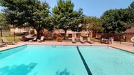 This outdoor amenity space centers around a generously sized swimming pool with clear, turquoise water and a classic rectangular design accented by a dark tile lane stripe. The pool deck is finished in a light-toned concrete surface, providing ample space for relaxation and movement around the perimeter. Multiple reclining lounge chairs and side tables are arranged along the poolside, offering comfortable spots to unwind and enjoy the surroundings. The area is enclosed by a sturdy, decorative wrought iron fence that adds a refined aesthetic while ensuring safety and privacy. Vibrant flowering plants, including splashes of red blooms, are positioned near the fence line, adding color and charm to the landscape. A canopy of large, mature trees lines the background, providing natural shade and a lush green backdrop that softens the open sky above. The overall ambiance is inviting and resort-like, combining clean architectural lines with verdant natural elements. Brick residential buildings are visible beyond the fencing, situating this pool area as a shared outdoor retreat within a well-maintained community environment. The space is bright and sun-drenched, ideal for outdoor leisure throughout the warmer months.
