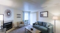This single-floor space centers around a welcoming living room designed for both comfort and functionality. The room is finished with plush carpet flooring and features neutral white walls that create a clean, airy backdrop. A large sliding glass door with vertical blinds, complemented by a nearby window dressed in light blue curtains, allows generous natural light to fill the space while offering outdoor access. The textured ceiling adds subtle architectural character throughout. The living area is well-proportioned, comfortably accommodating a blue-gray sofa accented with decorative throw pillows, a matching accent chair, and a rustic wooden coffee table. Wall-mounted shelving along one wall provides display space, while a framed decorative mirror adds depth and visual interest to the room. A small decorative console beneath the window serves as a charming focal point for potted plants, enhancing the inviting atmosphere. Overall, the space presents a bright, clean, and functional layout with a neutral color palette that lends itself well to a variety of interior styles.