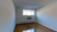 This single-floor space presents a straightforward and functional layout with a clean, minimalist aesthetic. The room features warm-toned hardwood floors with a glossy finish that adds a sense of richness and character to the otherwise neutral interior. Bright white walls provide a blank canvas, while the standard ceiling height contributes to a comfortable, enclosed atmosphere. A wide horizontal window fitted with white blinds allows natural light to filter into the space, brightening the room throughout the day. Climate control is addressed through a wall-mounted window air conditioning unit positioned directly beneath the window, paired with a baseboard heating system that runs along the exterior wall — providing year-round temperature regulation. White baseboards frame the perimeter of the room, adding a crisp, finished detail to the flooring transition. The overall ambiance is simple and unadorned, making the space highly adaptable for a variety of uses. The clean lines, practical heating and cooling infrastructure, and durable hardwood floors combine to create a no-frills interior that emphasizes utility and ease of use.