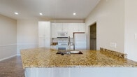 This single-floor residence offers a well-designed layout that balances functionality and comfort throughout its living spaces. The kitchen serves as a central highlight, showcasing warm golden granite countertops across a spacious peninsula with an undermount sink and pull-down faucet. White shaker-style cabinetry extends to the ceiling, providing generous storage, while white appliances — including a refrigerator, range, and built-in microwave — complete the space. An adjacent walk-in pantry offers additional storage convenience. The open living and dining areas feature durable hardwood-style flooring that runs throughout, complemented by crown molding, a ceiling fan, and multiple windows that invite abundant natural light. A distinctive natural wood beam accent adds architectural character to the transition area between rooms. The bedrooms are fitted with plush carpeting, ceiling fans, and windows with views to the outdoors, creating bright and comfortable retreats. The bathrooms maintain a clean, classic aesthetic with white tiled tub-shower combinations and chrome fixtures. Overall, the space presents a cohesive, neutral palette with warm wood tones and bright white finishes that create an inviting, move-in-ready atmosphere.