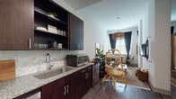 This thoughtfully designed single-floor suite blends bold aesthetics with functional living. The open-concept layout seamlessly connects a well-appointed kitchenette to a comfortable living and dining area. The kitchenette features rich dark wood cabinetry, granite countertops, a decorative tile backsplash, a stainless steel sink, microwave, and dishwasher — offering ample storage and everyday convenience. The living area is anchored by a brown leather sofa accented with vibrant throw pillows, complemented by framed wall art and a woven area rug. A small bistro-style dining nook sits beside tall windows dressed in dark curtains, allowing for abundant natural light while maintaining privacy. The bedroom is the undeniable focal point of the space, featuring a stunning floor-to-ceiling tropical botanical mural that envelops the room in lush, monochromatic grandeur. A woven rattan four-poster bed sits at the center, flanked by dark wood nightstands and finished with layered textiles, including a richly patterned kilim rug. The bathroom is clean and spacious, with wood-look flooring, granite-topped dark vanity, a tub-shower combination, and neutral tones throughout. Dark hardwood-style flooring flows consistently across the entire space, tying together the suite's warm, globally inspired character.