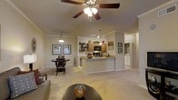 This single-floor residence showcases an open-concept layout that seamlessly connects the living, dining, and kitchen areas into one cohesive, flowing space. The living area is anchored by plush carpeted flooring and features a ceiling fan with integrated lighting, contributing to a comfortable and well-ventilated atmosphere. Crown molding along the upper walls adds a refined architectural detail throughout the space.

The kitchen is positioned centrally and is defined by a peninsula-style counter with a light granite surface, providing both a functional workspace and an informal dining or serving area. Warm wood-toned cabinetry, a built-in microwave, and open display shelving complete the kitchen's practical yet inviting design. A pendant light above the peninsula adds focused task lighting.

Adjacent to the kitchen, a cozy dining nook accommodates a small round table and chairs beneath a pendant light fixture, creating an intimate setting for meals. Framed artwork decorates the walls, adding character to the space. A glimpse of a laundry area with a washer is visible beyond the kitchen, indicating added in-unit utility. The warm, neutral color palette throughout gives the space a welcoming and cohesive ambiance.