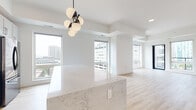 This single-floor residence showcases a contemporary open-concept design finished with light hardwood flooring and crisp white walls throughout. Large floor-to-ceiling windows frame sweeping urban skyline views, flooding the living and dining areas with abundant natural light. A striking multi-globe pendant chandelier anchors the kitchen island, which features an expansive marble-look quartz countertop that serves as both a functional workspace and a visual centerpiece.

The kitchen is outfitted with two-tone cabinetry—dark lower cabinets paired with light upper cabinets—complemented by a classic white subway tile backsplash, stainless steel Samsung appliances including a gas range with a wall-mounted hood, and a deep utility sink. Adjacent to the kitchen, the open living area is generously proportioned with multiple windows overlooking the cityscape.

The primary bathroom features a dual-sink vanity with dark cabinetry, white quartz countertops, matte black hardware, backlit mirrors, and a herringbone-tiled walk-in shower. A spacious walk-in closet with built-in shelving and hanging rods provides ample storage. Additional highlights include a dedicated in-unit laundry room with a full-size front-loading washer and dryer, and well-proportioned secondary rooms with city-facing windows.