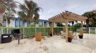 This single-story outdoor space presents a well-appointed patio area designed for relaxed outdoor enjoyment in a tropical setting. The centerpiece of the space is a sturdy wooden pergola with an open-slat roof, providing partial shade while maintaining an open, airy atmosphere. Beneath the pergola, wooden chairs and seating are arranged to create a comfortable gathering spot suited for leisure or casual socializing.

The patio surface is finished in light-toned concrete, extending generously across the outdoor area and bordered by a metal railing that defines the perimeter. Lush green hedging lines the fence, contributing to a sense of privacy and adding vibrant, natural color to the setting. Tall, mature palm trees rise prominently throughout the space, reinforcing the unmistakable tropical character of the environment and providing natural visual interest against the open sky.

Potted plants are thoughtfully placed around the pergola, adding layered greenery at ground level. A wood-toned waste receptacle is conveniently positioned along the railing. In the background, a multi-story building with blue accent trim and balconies is visible, providing context for the communal nature of the outdoor area. Overall, the space offers a casual, resort-like ambiance well-suited to an outdoor lifestyle.