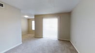 This single-floor space features an open layout with a calm, neutral aesthetic throughout. The main living area is finished with wall-to-wall beige carpeting, providing a soft and comfortable underfoot feel that extends into the adjoining room. Walls are painted in a warm, understated beige tone that complements the light-colored flooring and contributes to a cohesive, airy atmosphere.

A prominent sliding glass door serves as the focal point of the living area, dressed with full-length vertical blinds that allow for adjustable light control. When open, the blinds invite natural light to fill the space, enhancing its open feel. A secondary window in the adjacent room further supports natural illumination throughout.

The textured popcorn ceiling adds a traditional character to the interior, while clean white baseboards frame each room with a crisp finishing detail. The open sightline between the main living area and the connecting room creates a sense of continuity and spaciousness, making the layout feel practical and easy to navigate. Overall, the space presents a clean, move-in ready environment with a simple and functional design.