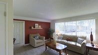 This single-story space centers around a welcoming living room that combines warmth and functionality. The most striking design element is the deep red accent wall, which creates a bold focal point and adds rich character to the room. A floating wood shelf on the accent wall provides a spot for decorative items, adding a personal touch to the space.

The living room is carpeted and comfortably sized, accommodating a cushioned sofa with decorative pillows, a matching armchair, and a wooden coffee table at the center. A floor lamp and a vase of white flowers contribute to the room's warm, inviting ambiance.

Natural light fills the space through a large window dressed with vertical blinds, offering a view of outdoor greenery. The textured popcorn ceiling adds a classic touch overhead, while clean white trim frames the walls and doorways throughout.

Overall, the space presents a cozy and functional layout with thoughtful design details, including the contrast between the rich accent wall and the neutral tones of the furnishings and carpet, creating a balanced and livable atmosphere.