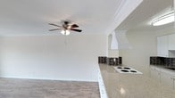 This single-floor space offers a clean, open-concept layout that seamlessly connects the living area to the kitchen, creating a bright and functional environment. The living area is finished with light hardwood-style flooring that adds warmth and a sense of spaciousness throughout. Crown molding runs along the ceiling, lending a refined architectural touch to the otherwise understated design. A ceiling fan with integrated lighting is centrally positioned, providing both air circulation and ambient illumination.

The kitchen is defined by a peninsula-style countertop with a dark surface that creates a striking contrast against the white cabinetry above. A classic dark mosaic tile backsplash adds texture and visual interest behind the cooking area. The kitchen is equipped with stainless steel appliances, including a full-size refrigerator. White upper cabinets provide ample storage while keeping the space feeling open and airy.

The neutral, warm-toned walls throughout the space complement the light flooring and white trim, contributing to a cohesive and inviting atmosphere. The open layout and thoughtful design details make this a practical and comfortable single-floor living space.