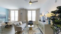 This single-floor residence at Overture offers a thoughtfully designed layout that balances functionality and comfort. A wide entryway corridor sets the tone upon entry, flanked by panel-style closet doors with sleek brushed nickel hardware, providing generous built-in storage. Dark hardwood-style flooring runs throughout, creating a warm contrast against the crisp white walls and trim.

The entryway opens into a well-proportioned living room bathed in natural light from multiple large windows dressed with elegant floor-length curtains. A ceiling fan with a brushed metal finish anchors the room from above, adding both style and practicality. The living space comfortably accommodates a sofa, accent chair, and coffee table arrangement centered on a decorative area rug, lending a relaxed yet polished atmosphere.

Along one wall, a sideboard credenza serves as an entertainment and décor focal point, complemented by accent lighting and mirrors. Lush greenery from an indoor plant adds a natural touch. The overall aesthetic is clean and contemporary, with a neutral palette of whites, grays, and soft blues that creates a cohesive, inviting environment throughout the space.