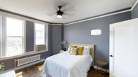 This single-floor space showcases a thoughtfully designed bedroom that blends classic architectural character with modern comfort. The room is dressed in cool, sophisticated grey walls accented by a crisp white crown molding border that adds a refined, traditional touch. Rich hardwood flooring grounds the space warmly, complementing the neutral color palette throughout.

A ceiling fan with an integrated light fixture serves as a functional centerpiece overhead, while a stylish arc floor lamp provides ambient lighting near the bed. The bedroom is furnished with a queen-sized bed dressed in crisp white bedding and accented with golden-yellow and patterned throw pillows, adding a warm pop of color against the cool grey backdrop.

Natural light filters in through a double-hung window, which also features a wall-mounted air conditioning unit and a classic radiator beneath it — details that speak to the space's enduring character. A compact nightstand with a floral arrangement adds a delicate, decorative touch, while a small side table rounds out the furnishings.

Overall, the space exudes a calm, welcoming ambiance, balancing timeless architectural elements with comfortable, contemporary styling that creates an inviting and restful environment.