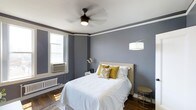 This single-floor space showcases a thoughtfully designed bedroom that blends classic architectural character with modern comfort. The room is dressed in cool, sophisticated grey walls accented by a crisp white crown molding border that adds a refined, traditional touch. Rich hardwood flooring grounds the space warmly, complementing the neutral color palette throughout.

A ceiling fan with an integrated light fixture serves as a functional centerpiece overhead, while a stylish arc floor lamp provides ambient lighting near the bed. The bedroom is furnished with a queen-sized bed dressed in crisp white bedding and accented with golden-yellow and patterned throw pillows, adding a warm pop of color against the cool grey backdrop.

Natural light filters in through a double-hung window, which also features a wall-mounted air conditioning unit and a classic radiator beneath it — details that speak to the space's enduring character. A compact nightstand with a floral arrangement adds a delicate, decorative touch, while a small side table rounds out the furnishings.

Overall, the space exudes a calm, welcoming ambiance, balancing timeless architectural elements with comfortable, contemporary styling that creates an inviting and restful environment.