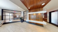 The Arlington 360 lobby presents a polished, contemporary entrance that sets a refined tone for the building. The focal point is a prominent reception desk clad in warm vertical wood paneling, prominently displaying the Arlington 360 branding in teal lettering. Above, a striking slatted wood ceiling adds warmth and architectural character, contrasting elegantly with the crisp white walls and light-toned large-format tile flooring that runs throughout the space.

The entry vestibule features dark-framed glass doors that invite natural light inward, flanked by a sleek wooden credenza topped with matching table lamps, creating a welcoming transition from exterior to interior. Recessed lighting and pendant fixtures provide balanced, ambient illumination throughout.

Adjacent to the main reception area, a glass-walled lounge space is visible, furnished with comfortable seating for residents and guests. Thoughtful design details, including two-tone wall treatments with subtle horizontal banding and dark base moldings, carry a consistent aesthetic through the corridors leading to restrooms and additional amenity spaces. A seating nook with a rounded accent chair and a circular wood table adds an intimate resting spot. Overall, the lobby delivers a cohesive, upscale ambiance that blends natural materials with modern finishes.