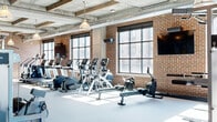 This expansive fitness center showcases a striking industrial aesthetic defined by exposed brick walls, coffered concrete ceilings with heavy timber beams, and oversized steel-framed windows that flood the space with abundant natural light and city views. The facility spans a generous single floor, thoughtfully organized into distinct training zones. The main exercise floor features a comprehensive array of Hammer Strength cable machines, power racks, and weight stations, complemented by a dedicated free weights area equipped with a full dumbbell rack and adjustable benches, all set against mirrored walls. The cardio zone offers an extensive lineup of treadmills, ellipticals, stair climbers, and rowing machines positioned along the windowed perimeter. A separate multi-purpose studio features warm hardwood flooring, floor-to-ceiling mirrors, wall-mounted flat-screen TVs, and ample open space suitable for yoga, cycling, or group fitness classes. Pendant industrial lighting hangs throughout, reinforcing the space's refined yet functional character. Exposed ductwork and concrete surfaces complete the modern industrial look, creating an energizing and well-equipped environment designed to support a full range of fitness activities.
