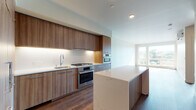 This contemporary single-floor residence showcases a thoughtfully designed open-concept layout that seamlessly connects the kitchen and living areas. The kitchen is a standout feature, outfitted with flat-panel walnut-toned cabinetry, stainless steel appliances including a gas cooktop, double wall ovens, and a full-size range. A generous island with a striking marble-look quartz countertop anchors the space, offering both additional prep space and a casual gathering spot. Recessed lighting and under-cabinet illumination add a polished finish.

Rich, warm-toned hardwood flooring flows throughout the open living area, which is bathed in natural light through large floor-to-ceiling windows that frame impressive views of the surrounding cityscape and treetops. A wall-mounted mini-split system ensures year-round comfort.

The bedroom benefits from the same expansive windows and sunlit ambiance, complemented by a spacious walk-in closet outfitted with adjustable wire shelving and hardwood flooring. The bathroom features large-format linear tile in neutral tones wrapping a full soaking tub and shower combination.

A dedicated in-unit laundry area houses side-by-side front-load washer and dryer units on pedestals, completing this well-appointed residence.