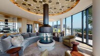 This sophisticated lounge space is defined by its striking circular design language and high-end architectural details. At the heart of the room sits a dramatic suspended cylindrical fireplace with a glass surround and blue fire glass base, anchored beneath a decorative recessed ceiling medallion adorned with a gold-and-white organic patterned mosaic. Curved, velvet sectional sofas are arranged around this focal point, complemented by sleek armchairs and sculptural wooden accent tables, creating an intimate yet open gathering environment. Light wide-plank hardwood flooring flows throughout, lending warmth to the modern aesthetic. Floor-to-ceiling glass walls wrap around the space, flooding the interior with abundant natural light and framing expansive panoramic views of the surrounding cityscape and skyline. These glass walls open directly onto a generous outdoor terrace, furnished with seating and planters, seamlessly extending the living area outdoors. A separate dining area features a rounded table surrounded by cream upholstered chairs beneath an elegant capiz shell chandelier, paired with a built-in wet bar finished in warm wood cabinetry and marble backsplash. Slender white structural columns punctuate the perimeter without interrupting the openness of the layout, reinforcing the space's airy, elevated ambiance.