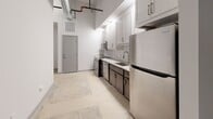 Located at 104 E Second St in Richmond, VA, this single-floor unit blends industrial character with modern finishes throughout. Polished concrete floors run continuously across the space, complementing soaring ceilings that showcase exposed black piping, ductwork, and conduit — hallmarks of an adaptive reuse aesthetic.

The kitchen is a functional highlight, featuring white shaker upper cabinets paired with dark lower cabinetry, a white subway tile backsplash, light countertops, and a full suite of stainless steel appliances including a refrigerator, range, microwave, and dishwasher. The generous galley-style layout provides ample prep and storage space, with elevated ceilings lending an open, airy feel even within the kitchen corridor.

The bathroom is cleanly finished with a full tub-shower combination surrounded by large-format white tile, brushed nickel fixtures, a toilet, and a vanity topped with dark stone countertops. Recessed lighting provides a bright, polished atmosphere.

A dedicated laundry closet houses a stacked washer and dryer unit, offering added convenience within the home. The main living area is expansive and open, with bright white walls and abundant room to customize the layout. Natural light filters in through windows, and the overall ambiance is modern and loft-inspired, well-suited for those who appreciate clean lines and industrial design elements.