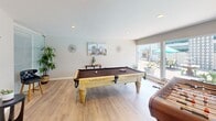Located at 2635 Milton Ave in Fullerton, California, this dedicated game room offers a well-appointed recreational retreat designed for leisure and entertainment. The single-floor space is finished with light wood-tone luxury vinyl plank flooring that lends a clean, modern feel throughout.

At the heart of the room sits a handsome full-size pool table with an ornately carved light wood frame and rich brown felt, complemented by a foosball table that adds to the variety of entertainment options. Recessed lighting overhead ensures the space is well-lit for gameplay, while the neutral warm-white walls provide a crisp, inviting backdrop.

A defining architectural feature is the expansive wall of sliding glass doors spanning nearly the full length of one side of the room, flooding the interior with natural light and providing direct views of a sparkling outdoor pool, sundeck, and patio area. Decorative lattice-style privacy screens on the adjacent exterior add a mid-century modern touch to the outdoor aesthetic.

A frosted glass sliding interior partition on one wall adds a contemporary design element while allowing light to pass through. Thoughtful touches such as potted plants, coastal-themed artwork, and starburst wall clocks complete the relaxed California ambiance, making this game room a standout communal amenity.