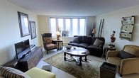 This single-floor space centers around a thoughtfully arranged living room that balances comfort and classic style. A wide, multi-pane window serves as the focal point of the room, drawing in abundant natural light and offering an open view of the exterior. The neutral, cream-toned walls complement the light-colored carpet, creating a bright and airy ambiance throughout.

The living room is furnished with a large, dark upholstered sectional sofa accented with decorative throw pillows, paired with a traditional patterned armchair that adds a touch of character. A ornate dark wood coffee table with carved legs anchors the seating area atop a muted, vintage-style area rug that adds warmth and texture to the space.

Additional furnishings include a dark wood sideboard, a decorative leaning ladder against the wall, and coordinating side tables with table lamps that contribute to the room's layered, classic aesthetic. Decorative elements such as framed wall art, dried branches in tall vases, and carefully placed accessories complete the inviting, well-curated atmosphere of this living space.